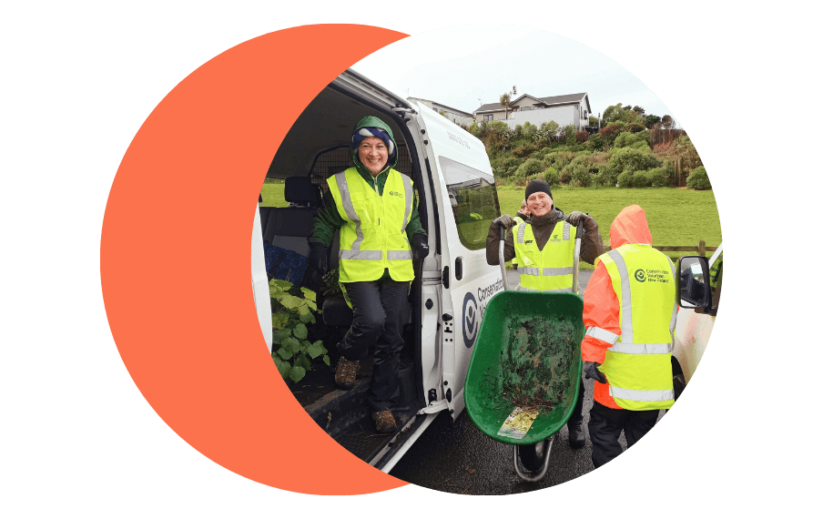 Heather and David from Thankyou Payroll wearing high vis, volunteer tree planting for Conservation Week. Heather stands in a truck and David is holding a wheel barrow. Both look at the camera smiling.